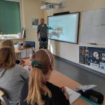 Uczennice w klasie słuchają prowadzącego, który pokazuje slajd na tablicy multimedialnej i napis "Budowa ogrodu deszczowego"