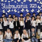 Na tle niebieskiej kotary z napisem- ‘Ślubowanie 2023”- stoi klasa 1b z wychowawczynią.