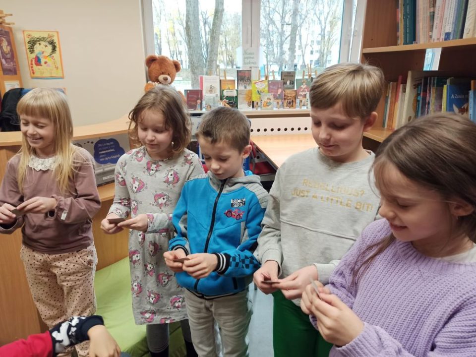 Pięcioro dzieci stoi na tle okna i regału z książkami. Dzieci z zamkniętymi oczami dotykają dłońmi karteczki, które trzymają.